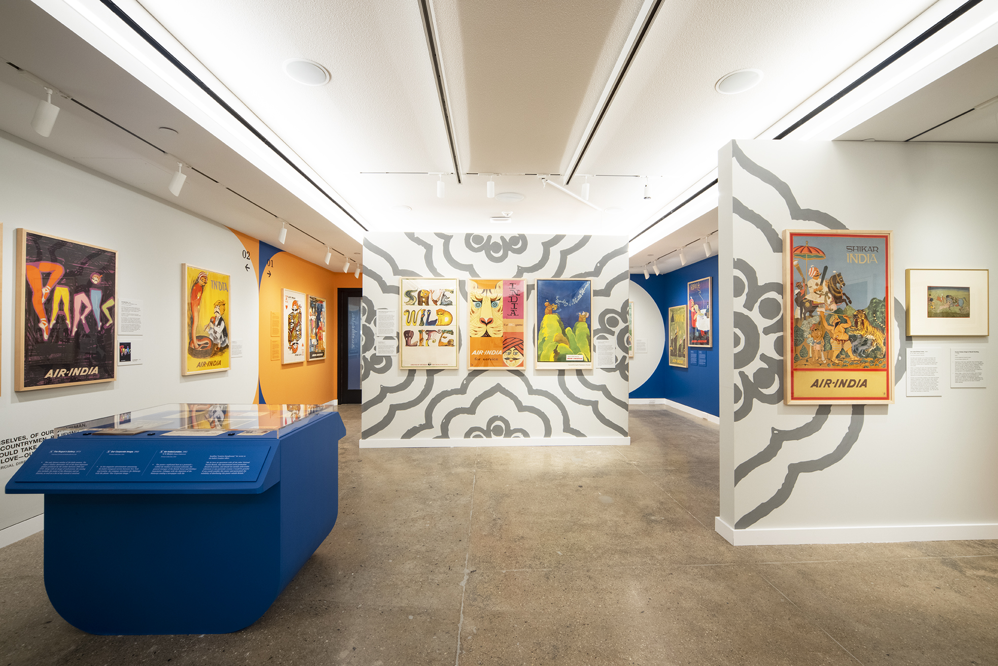 A gallery space with framed posters from Air India airlines lining the outer walls and a wall space in the center of the room. On the left there is a blue, rounded display case of printed Air India historical objects.