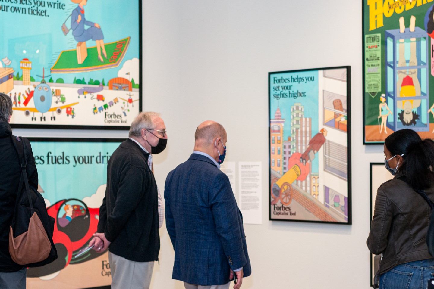 Two figures look at a poster on the wall surrounded by other posters and visitors.
