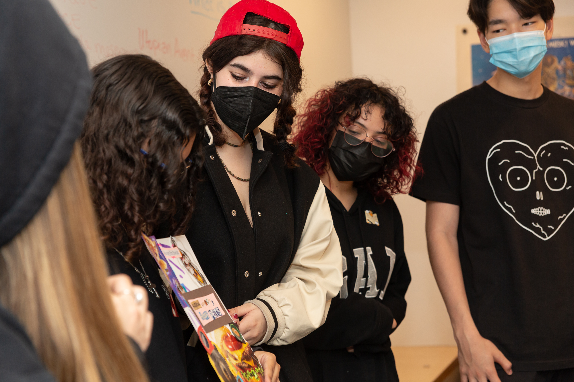 A group of students wearing face masks point at and observe a paper with collage work