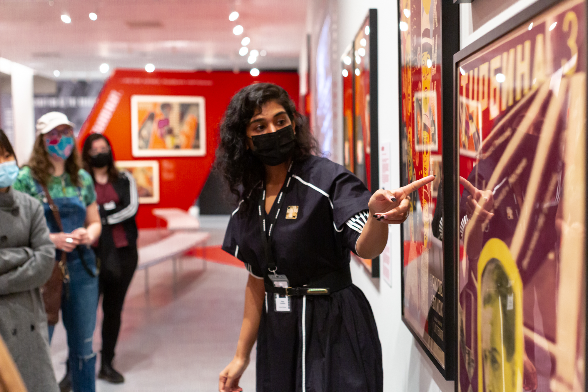 An educator wearing all black and a Poster House badge around her neck stands in front of students in a gallery and points at a framed poster hanging on a gallery wall
